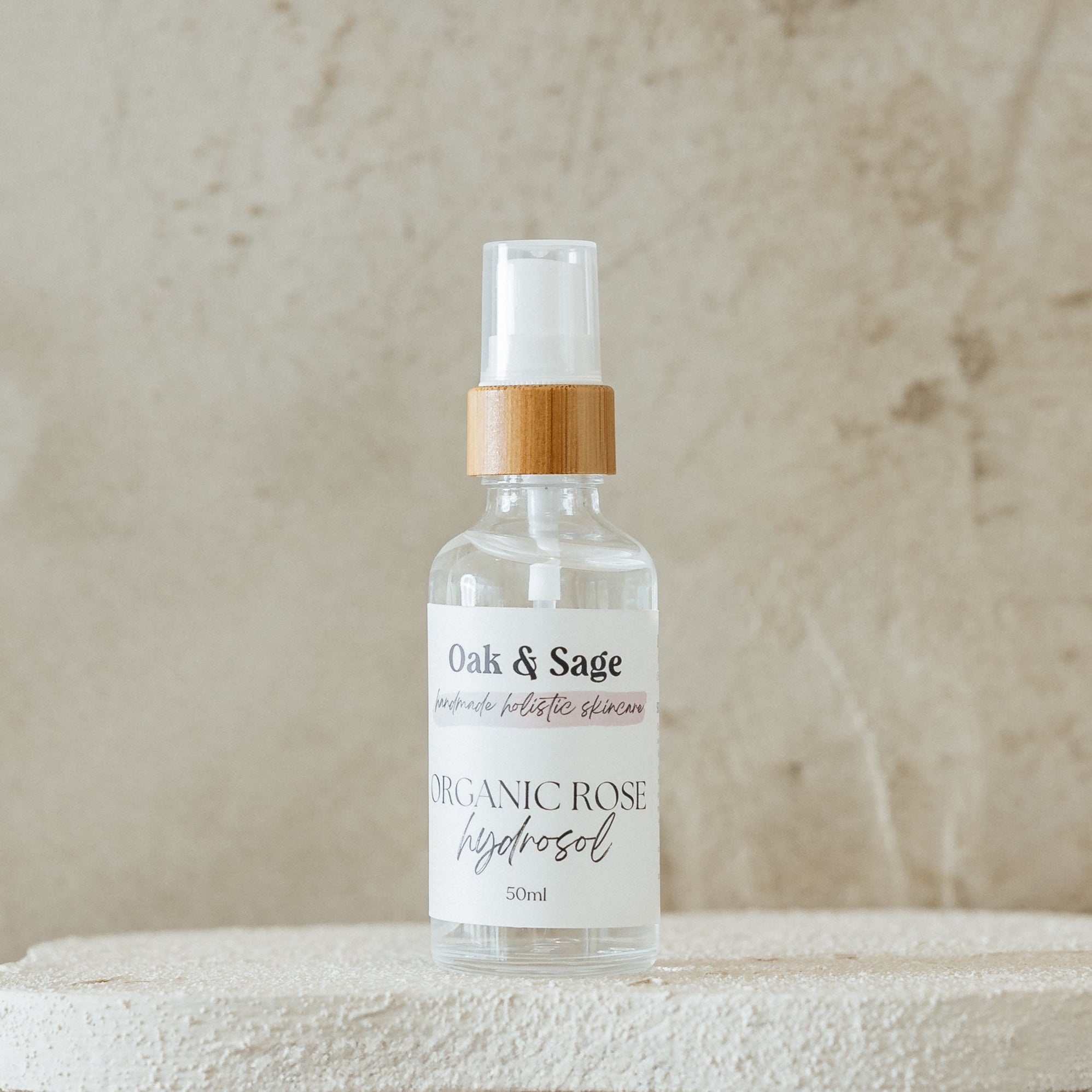 Oak & Sage Organic Calendula Hydrosol in glass spray bottle with bamboo top, displayed on white textured pedestal against warm marbled background.