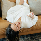 Relaxed woman in a white robe lying on a leather couch, holding Oak & Sage Peppermint Lip Balm, natural tallow-based skincare for soft, hydrated lips.