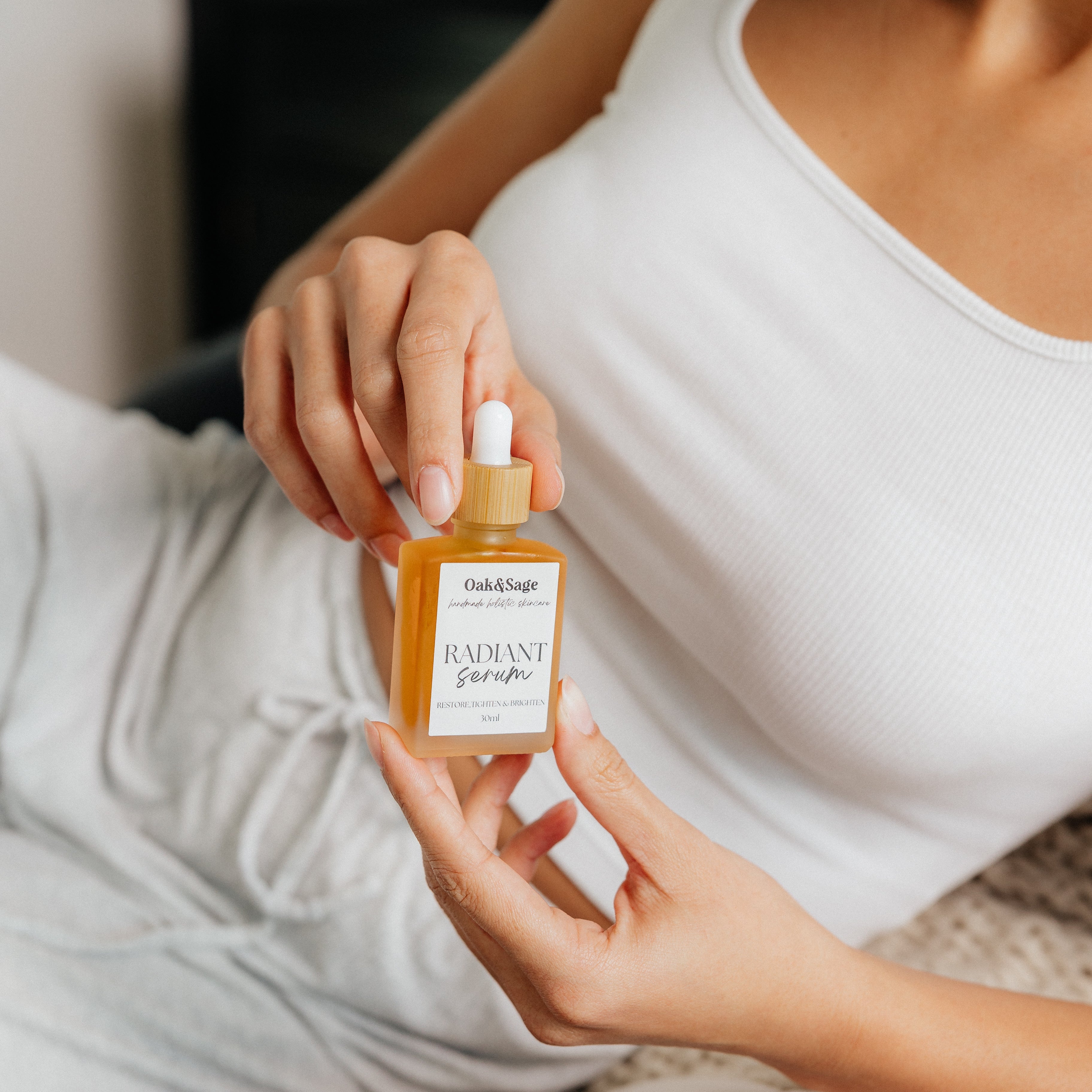 Woman in white tank top holding Oak & Sage Radiant Serum bottle, showcasing skincare product for restoring, tightening, and brightening skin.