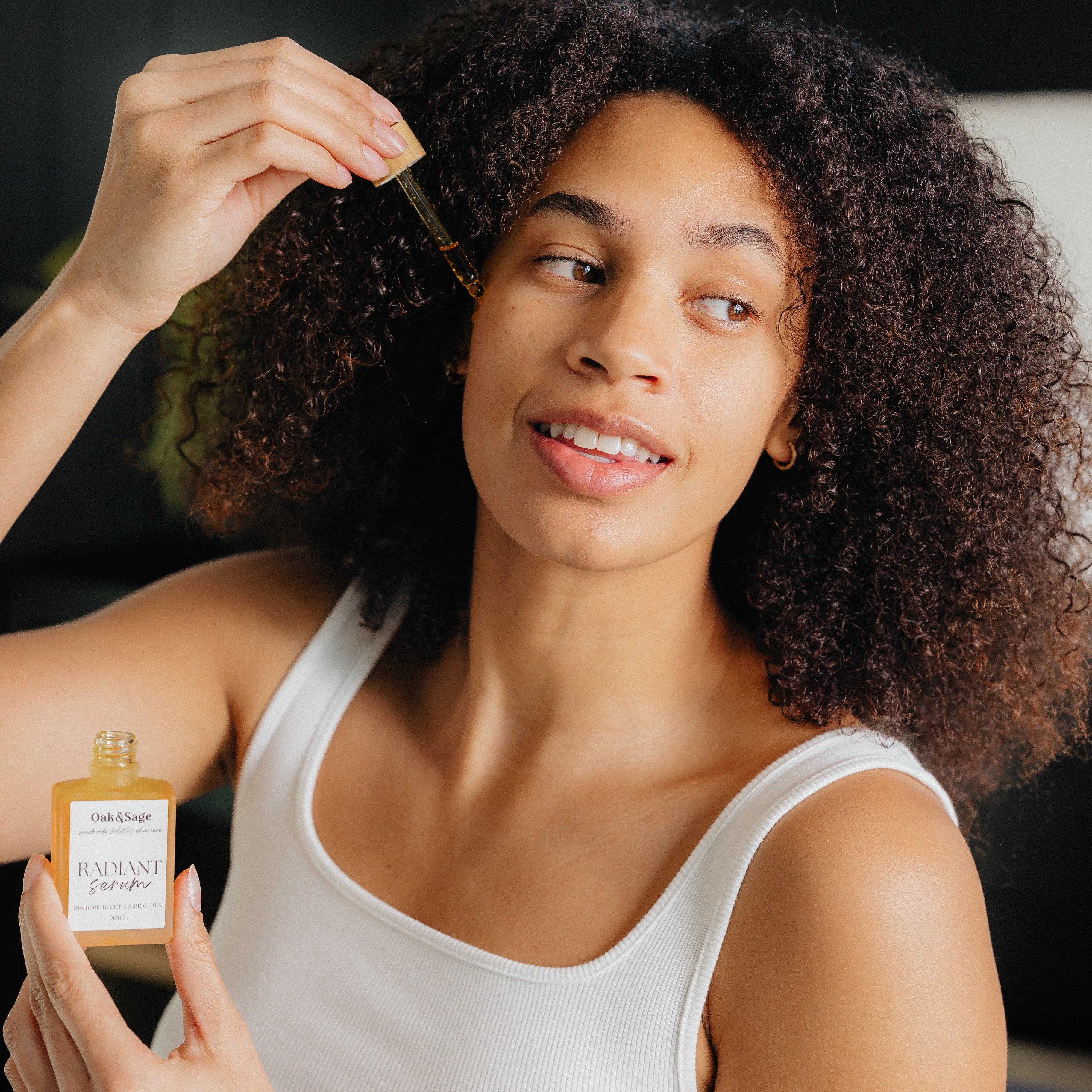 Smiling woman applying Oak & Sage Radiant Serum to her face using a dropper, demonstrating natural skincare routine for glowing, healthy skin.