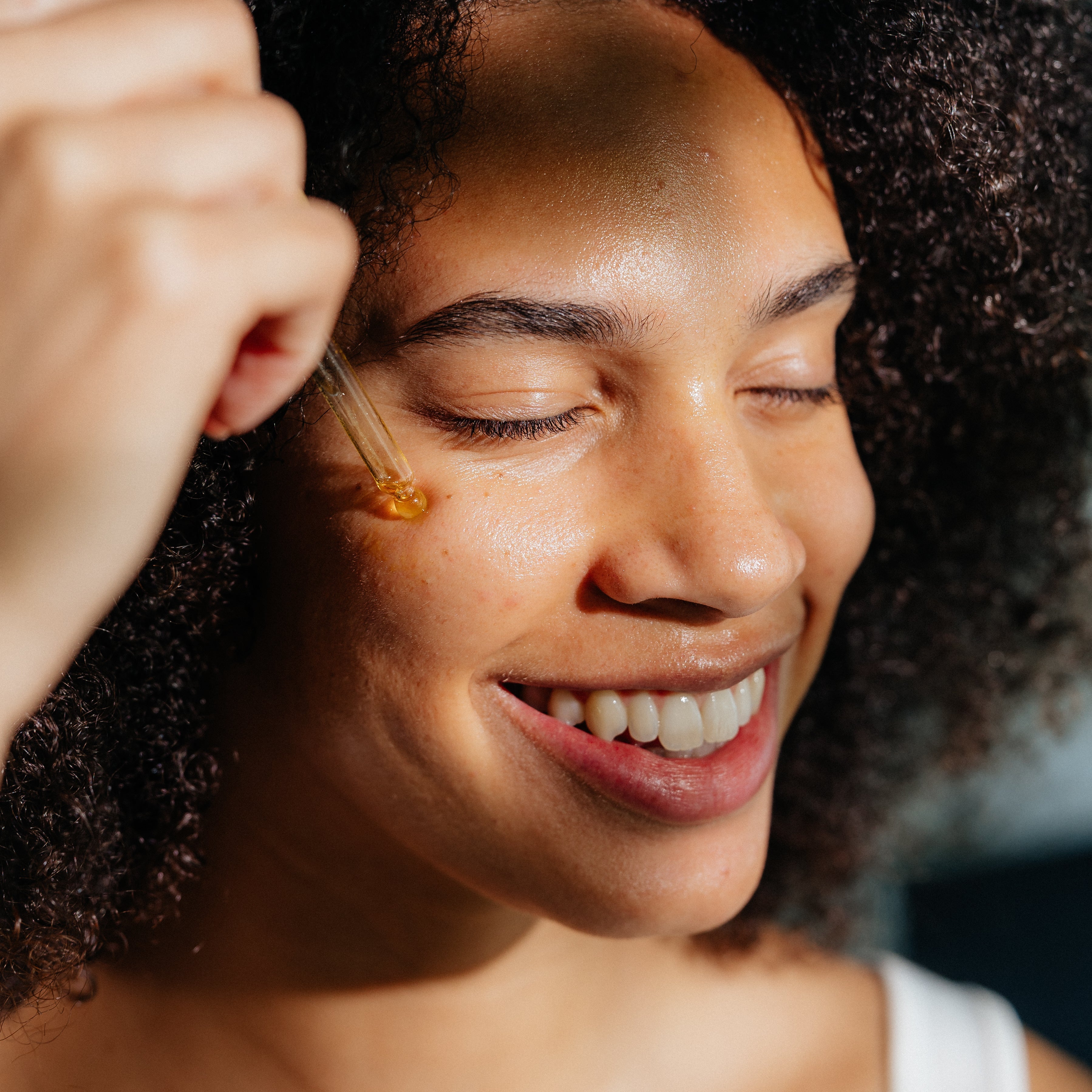 Smiling woman applying Oak & Sage Radiant Serum with dropper to cheek, highlighting natural glow and hydrating skincare benefits.