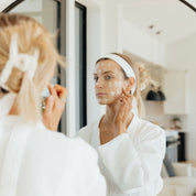Woman in white robe and headband applying Oak & Sage Youthful Cream in front of a mirror, skincare routine with white moisturizer dots on her face in a modern, bright bathroom.