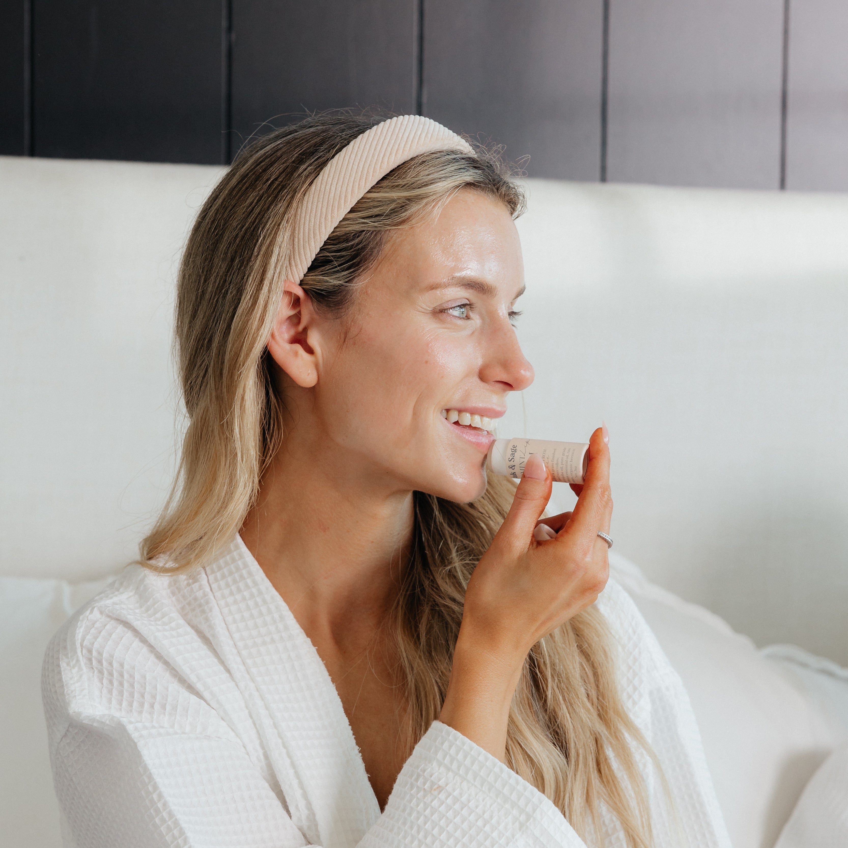 Woman in a white robe applying Oak & Sage Vanilla Lip Balm with a smile, highlighting natural skincare and eco-conscious beauty in a cozy morning routine.