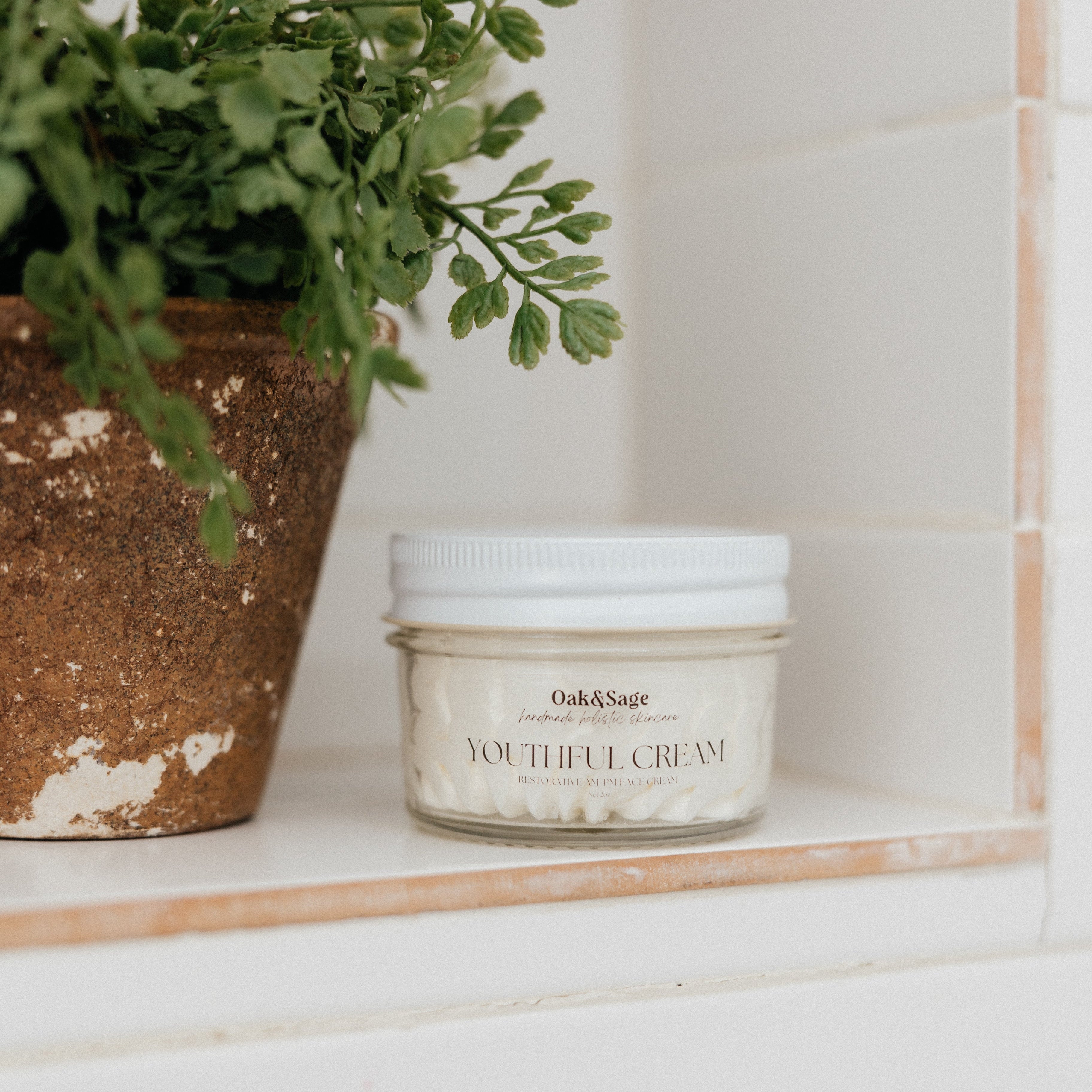 Oak & Sage Youthful Cream in a glass jar with white lid, displayed on a tiled shelf beside a rustic terracotta pot with green herbs, natural skincare aesthetic.