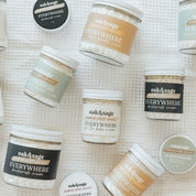 Flat lay of Oak & Sage Everywhere Cream collection featuring natural body butters in scents like Warm Vanilla, Backwoods, Eucalyptus Lavender, and For the Babe—handmade holistic skincare in reusable glass jars