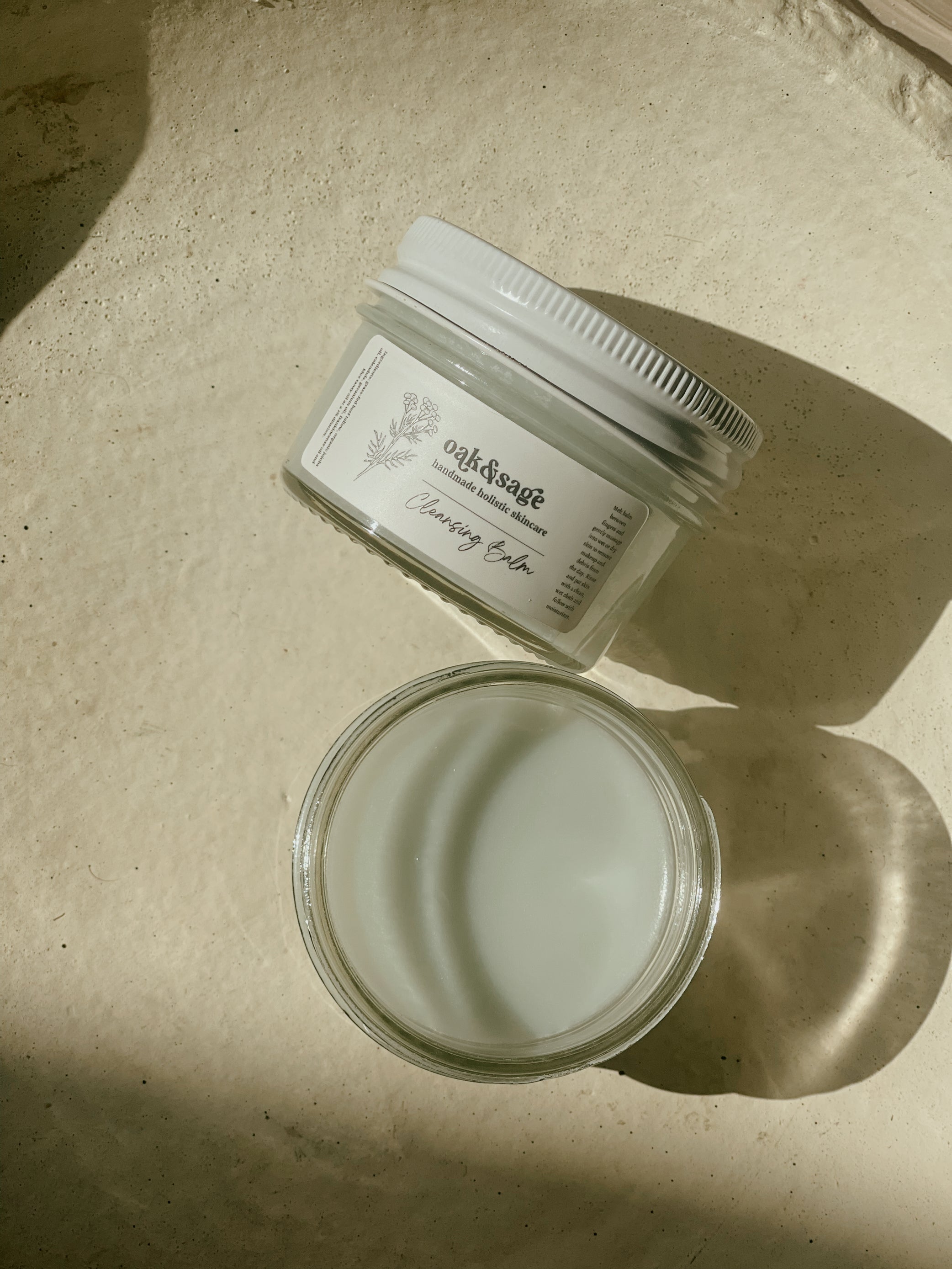Oak & Sage Cleansing Balm in open and closed glass jars, displayed in natural sunlight on a warm-toned surface — showcasing the balm’s creamy texture and artisanal skincare aesthetic.