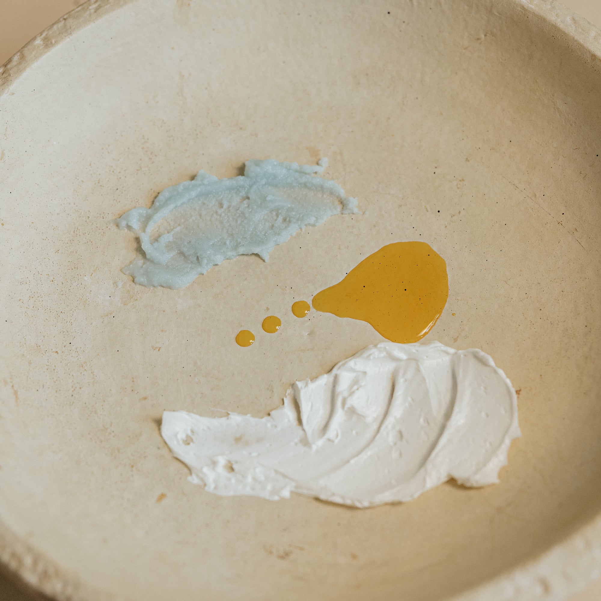 Textural swatches of Oak & Sage Complete Skincare Set showing blue Cleansing Balm, golden Radiant Serum, and whipped Youthful Cream on a neutral ceramic plate.