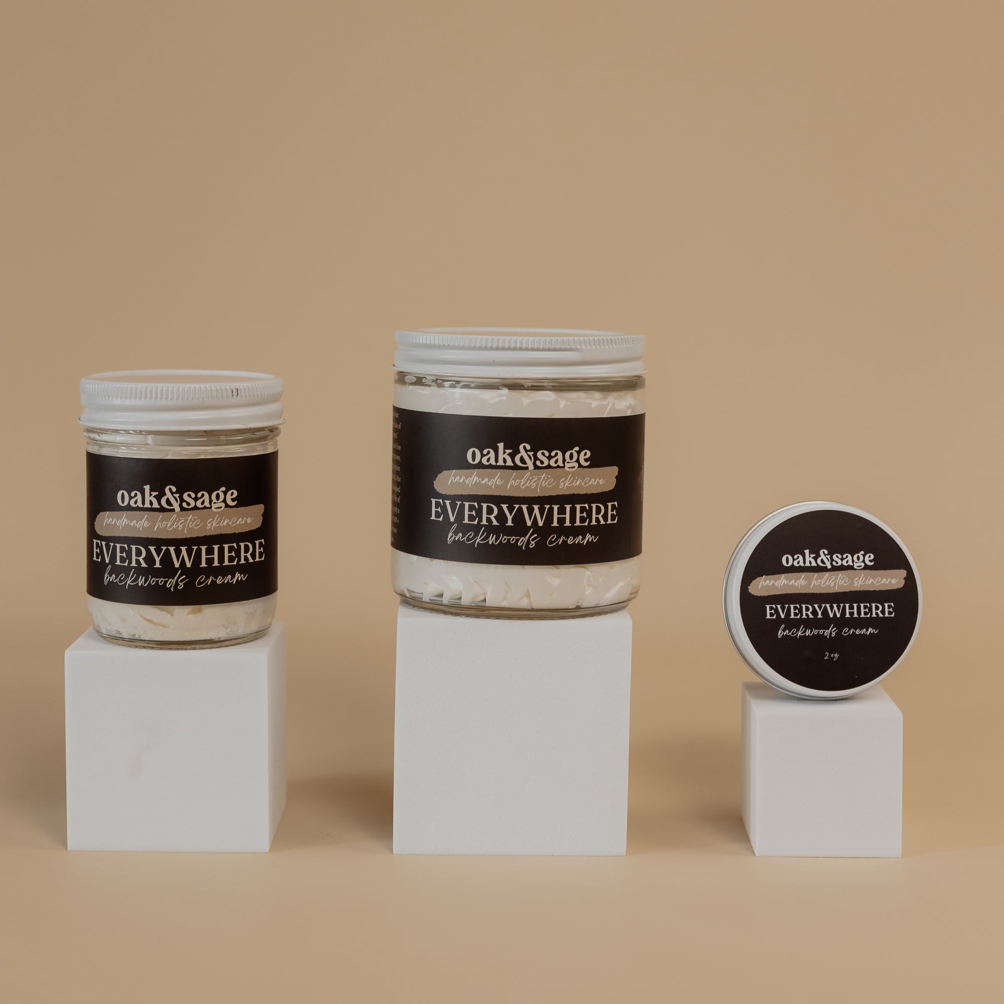 Oak & Sage Everywhere Cream in Backwoods scent displayed in three sizes—large jar, medium jar, and travel tin—on minimalist white pedestals against a warm beige background