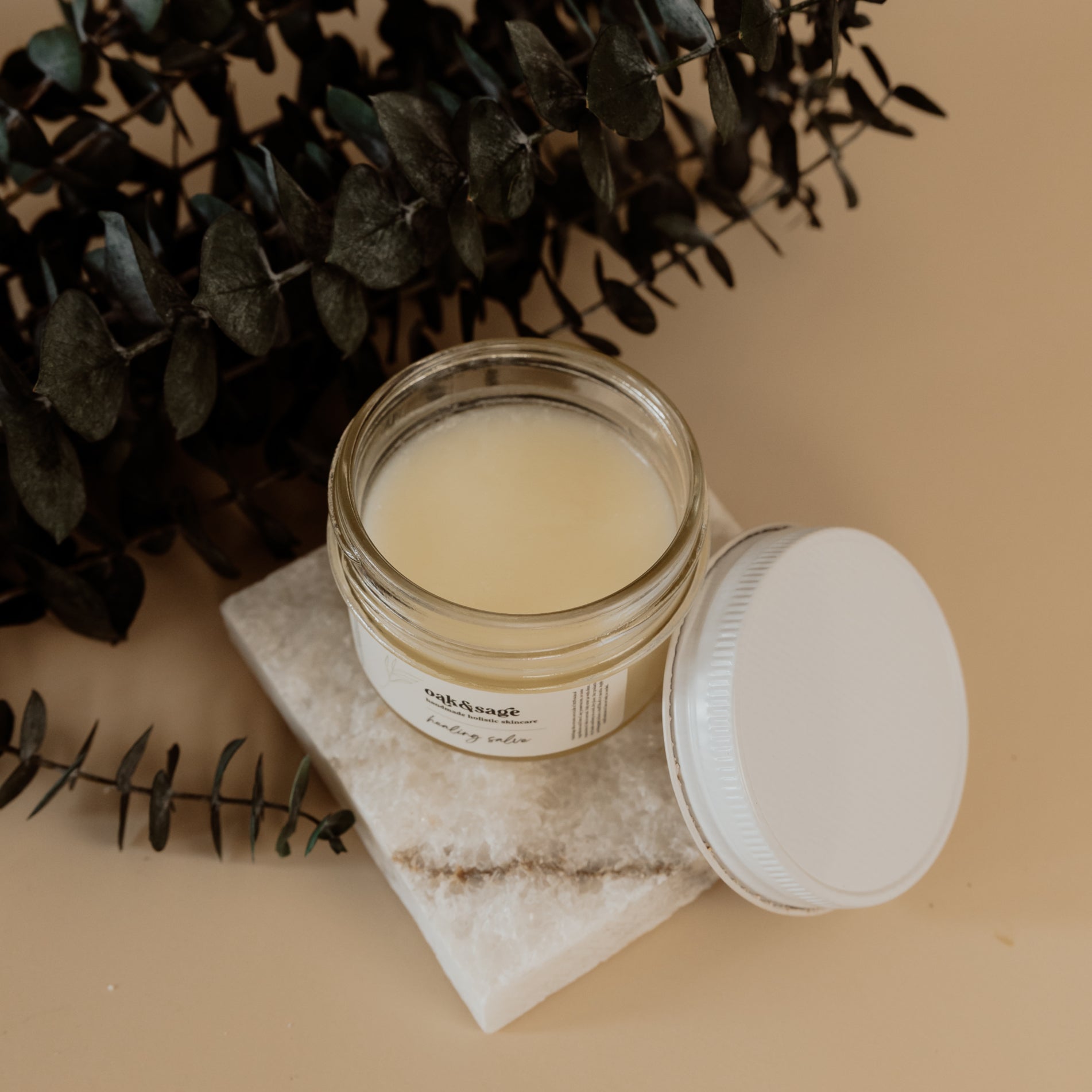 Open jar of Oak & Sage Healing Salve on marble tile with eucalyptus, showcasing creamy natural texture of tallow-based skin repair balm.