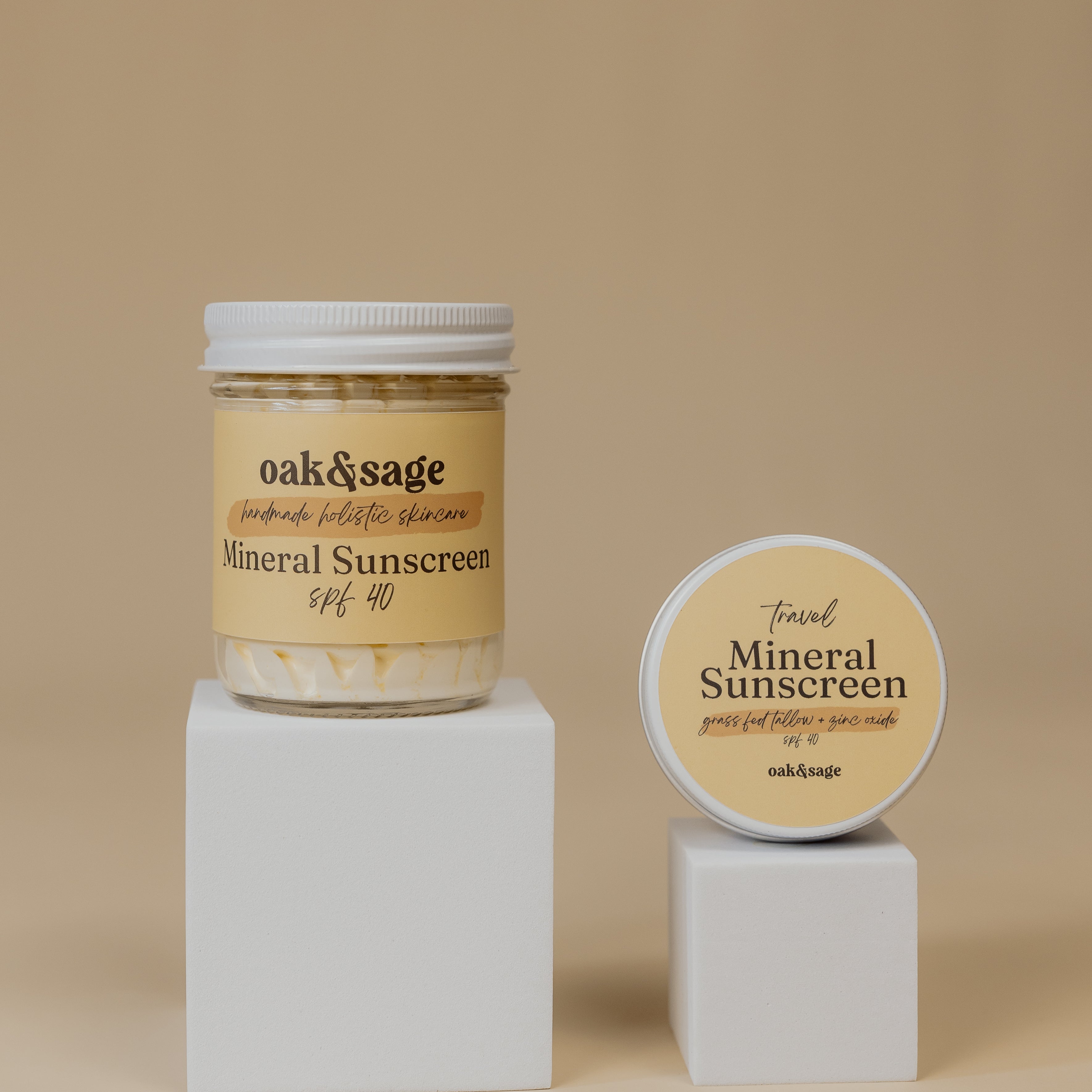 Oak & Sage Mineral Sunscreen SPF 40 in 8 oz glass jar and 2 oz travel tin, displayed on white pedestals against neutral beige background.
