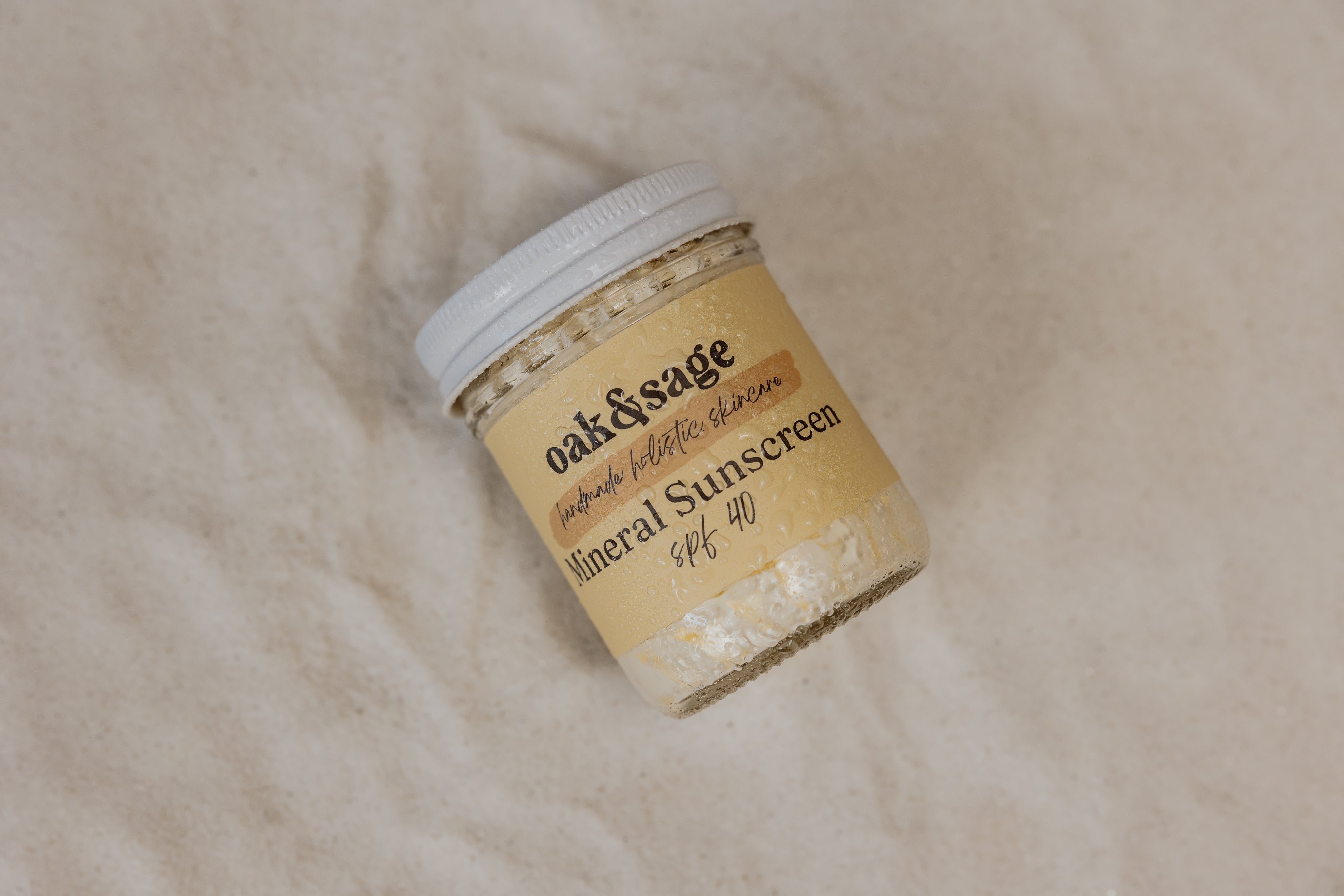 Oak & Sage Mineral Sunscreen SPF 40 in 8 oz glass jar with condensation, resting on soft sand to highlight beach-ready, natural sun protection.