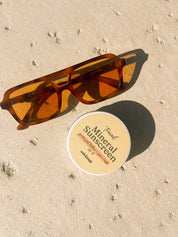 Oak & Sage Travel Mineral Sunscreen SPF 40 tin with grass-fed tallow and zinc oxide beside retro amber sunglasses on sunlit concrete surface.