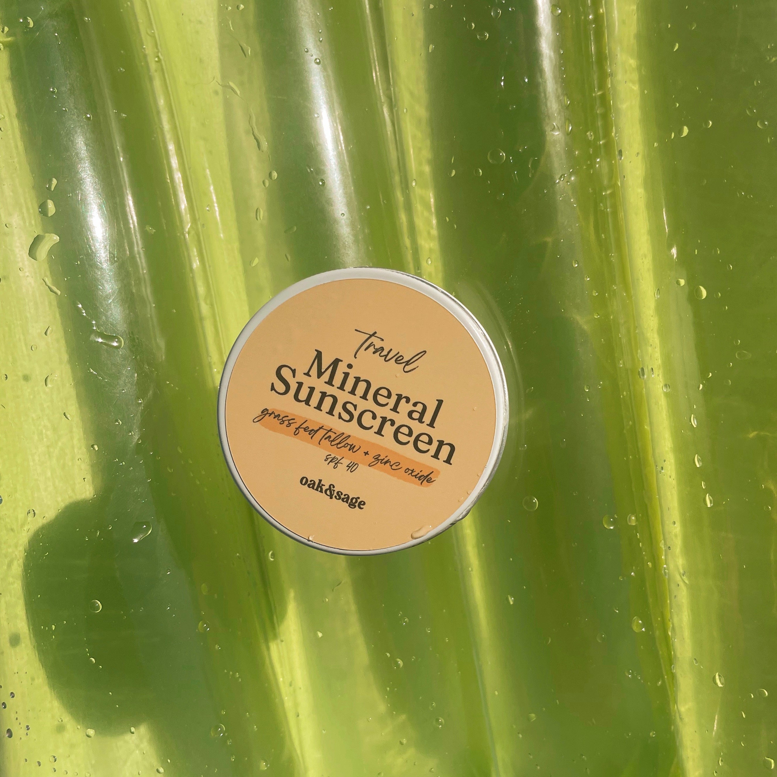 Oak & Sage Travel Mineral Sunscreen SPF 40 tin with grass-fed tallow and zinc oxide, resting on bright green pool float with water droplets.