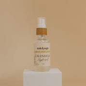 Oak & Sage Organic Calendula Hydrosol in glass spray bottle with bamboo top, displayed on white pedestal against warm beige background.