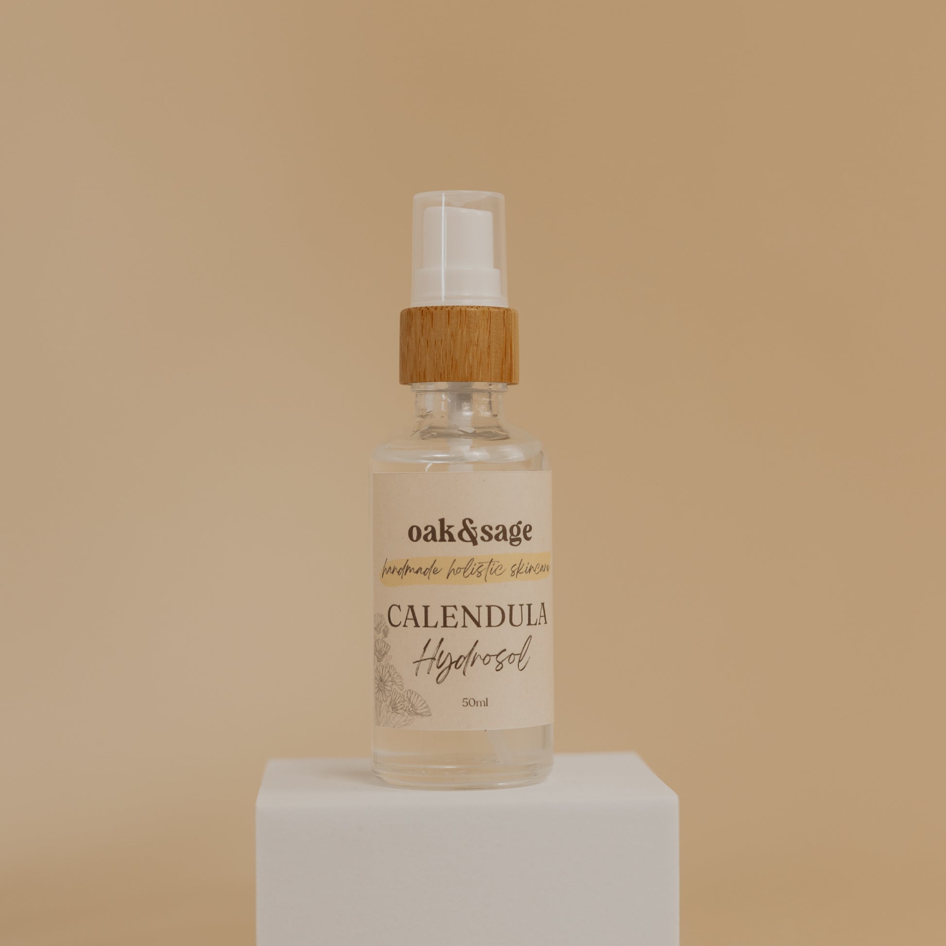 Oak & Sage Organic Calendula Hydrosol in glass spray bottle with bamboo top, displayed on white pedestal against warm beige background.