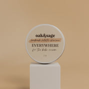 Oak & Sage For The Babe Everywhere Cream in a 2 oz travel-size tin, displayed on a modern white pedestal against a neutral beige background