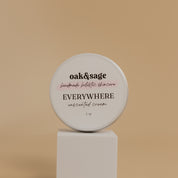 Oak & Sage Unscented Everywhere Cream in a 2 oz travel-size tin, displayed on a modern white pedestal against a neutral beige background