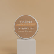 Oak & Sage Warm Vanilla Everywhere Cream in a 2 oz travel-size tin, displayed on a modern white pedestal against a neutral beige background