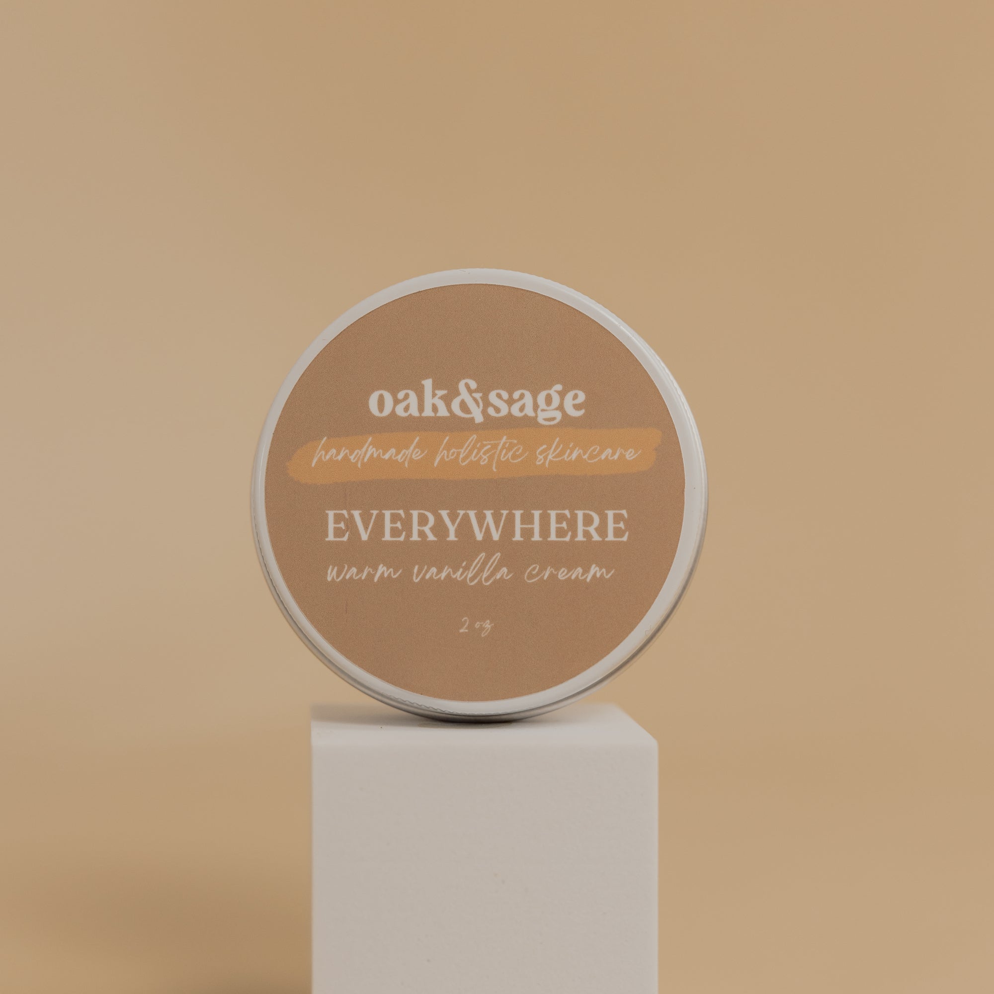 Oak & Sage Warm Vanilla Everywhere Cream in a 2 oz travel-size tin, displayed on a modern white pedestal against a neutral beige background