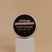 Oak & Sage Backwoods Everywhere Cream in a 2 oz travel-size tin, displayed on a modern white pedestal against a neutral beige background