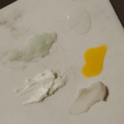 Close-up swatches of Oak & Sage Ultimate Skincare Set textures including Radiant Serum, Cleansing Balm, Exfoliating Balm, Youthful Cream, and Lip Balm on neutral ceramic surface.